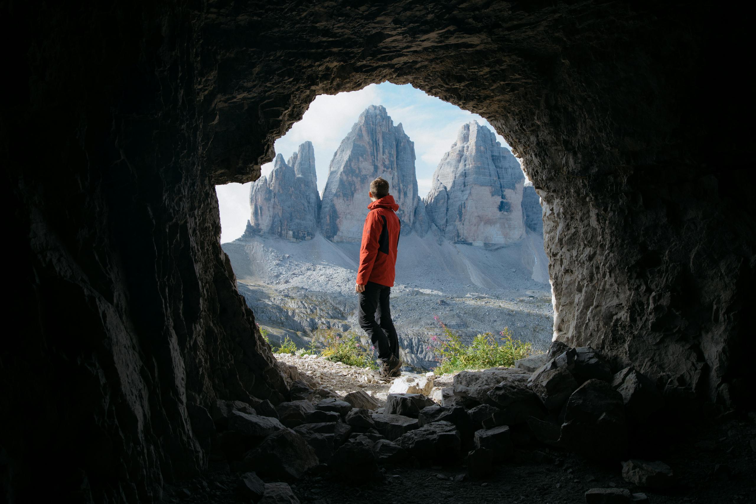 Man in red jacket standing in cave entrance, viewing Tre Cime di Lavaredo in Italy.